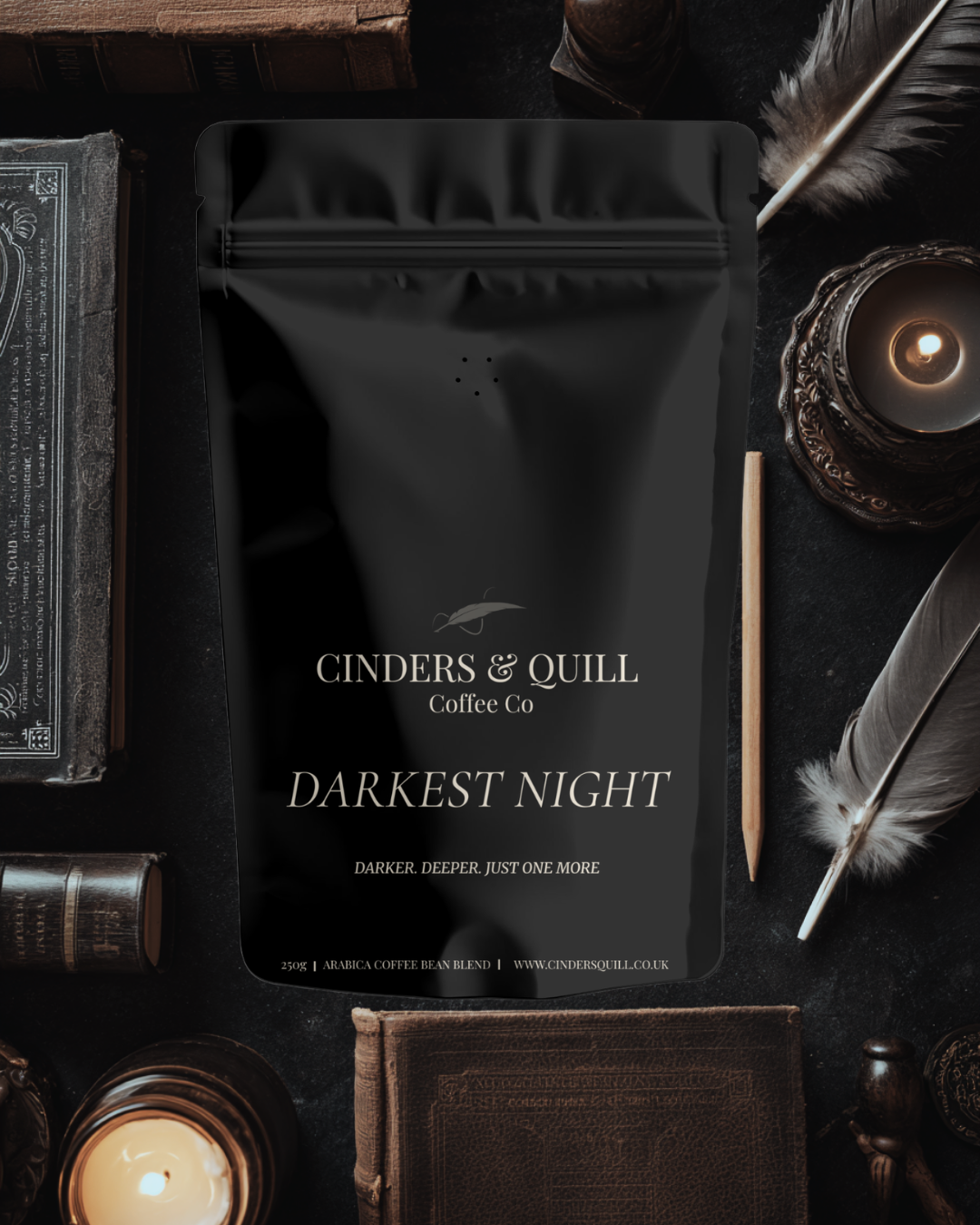 Black coffee bag labeled 'Cinders & Quill Coffee Co Darkest Night' on a dark surface with candles and feathers.